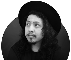 Foto de Pedro Henrique Moura Leite em preto e branco, utilizando um chapeu preto e uma camiseta preta, ele está com o cabelo solto e olhando levemente para o lado esquerdo.